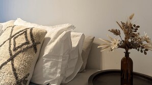 A bedroom with a bed, pillows, a round side table, and a vase with dried flowers.