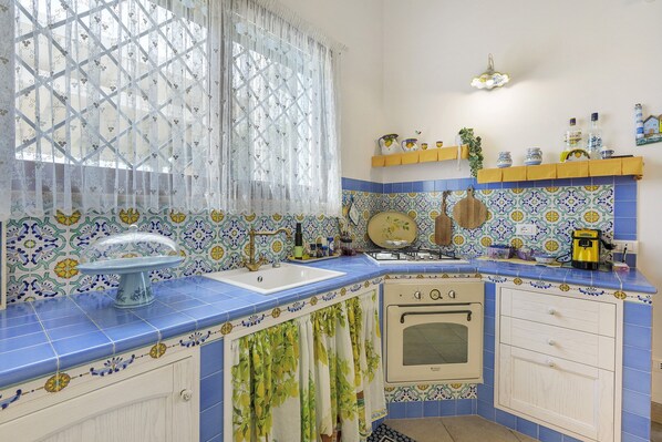 A kitchen with blue and white tiles, a sink, a stove, and a window with a sheer curtain. - Casa Babalù sea view with private pool (Partinico)