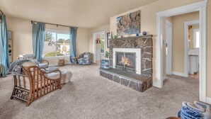 A spacious living room with a stone fireplace, a large window with curtains, and a cozy seating area with a wooden chair and a sofa. - Expansive Decks, Two Living Areas with Fireplaces! (Lincoln City)