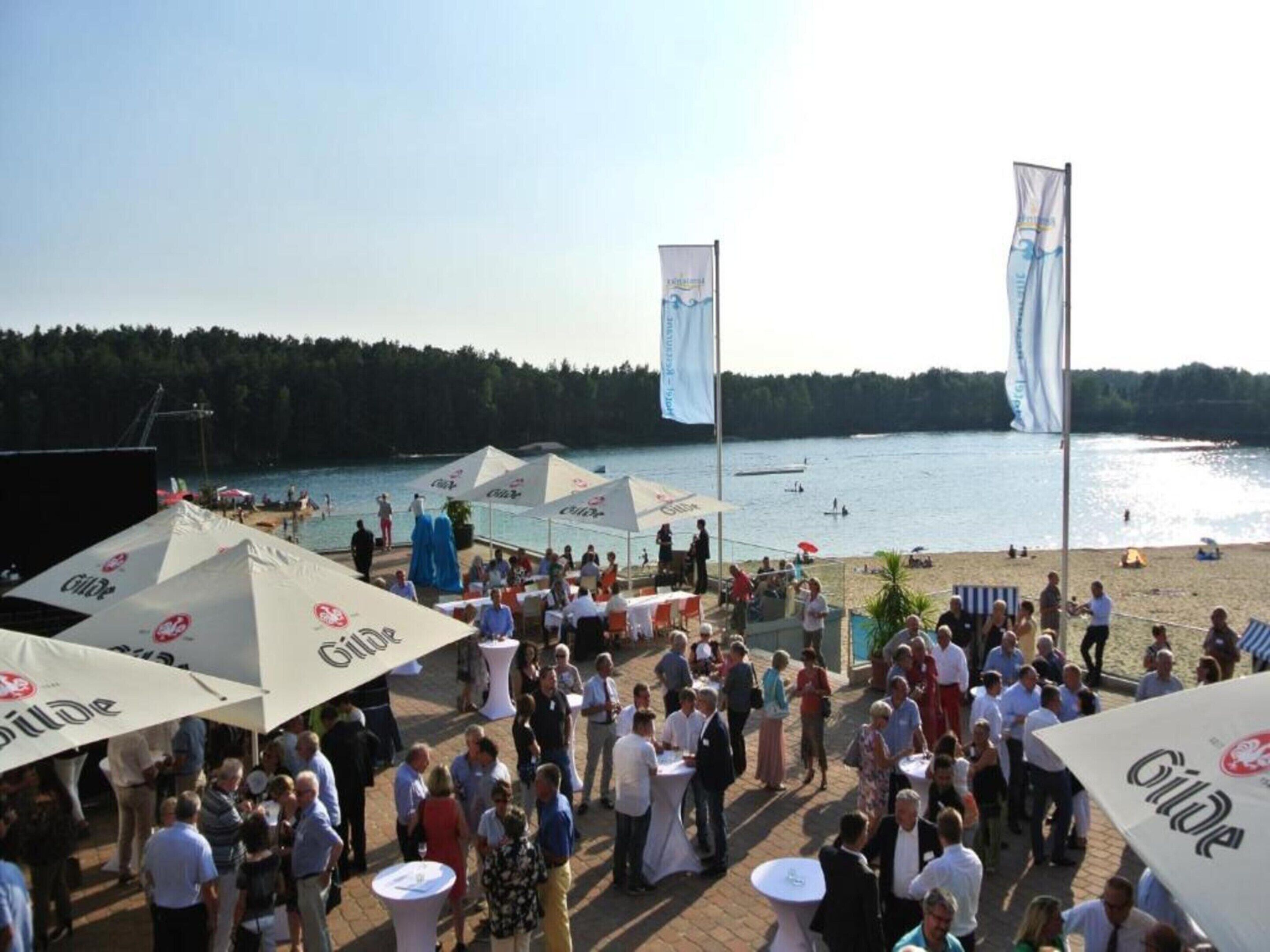 A crowded outdoor event with people gathered around tables under umbrellas, overlooking a lake with boats and a beach in the background.