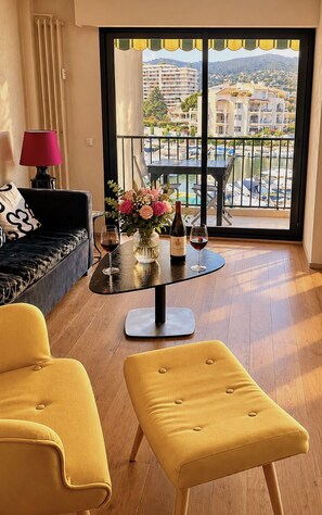 Un salon moderne avec un canapé, deux fauteuils jaunes, une table basse en verre surmontée d’un vase de fleurs et de bouteilles de vin, et une vue sur une marina et des immeubles, le tout visible à travers de grandes fenêtres.