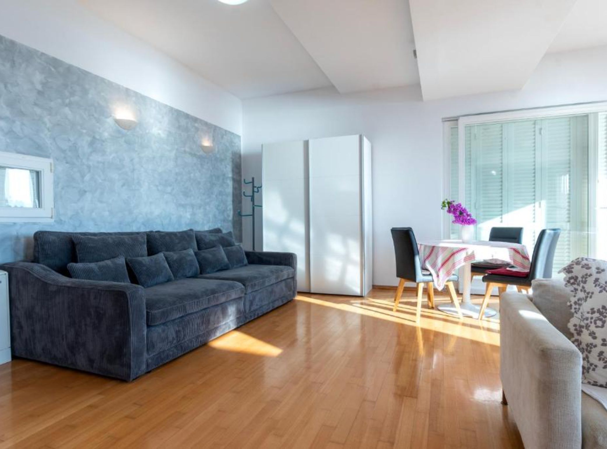 A modern living room with a grey sofa, wooden flooring, a dining table with chairs, and a window with blinds.