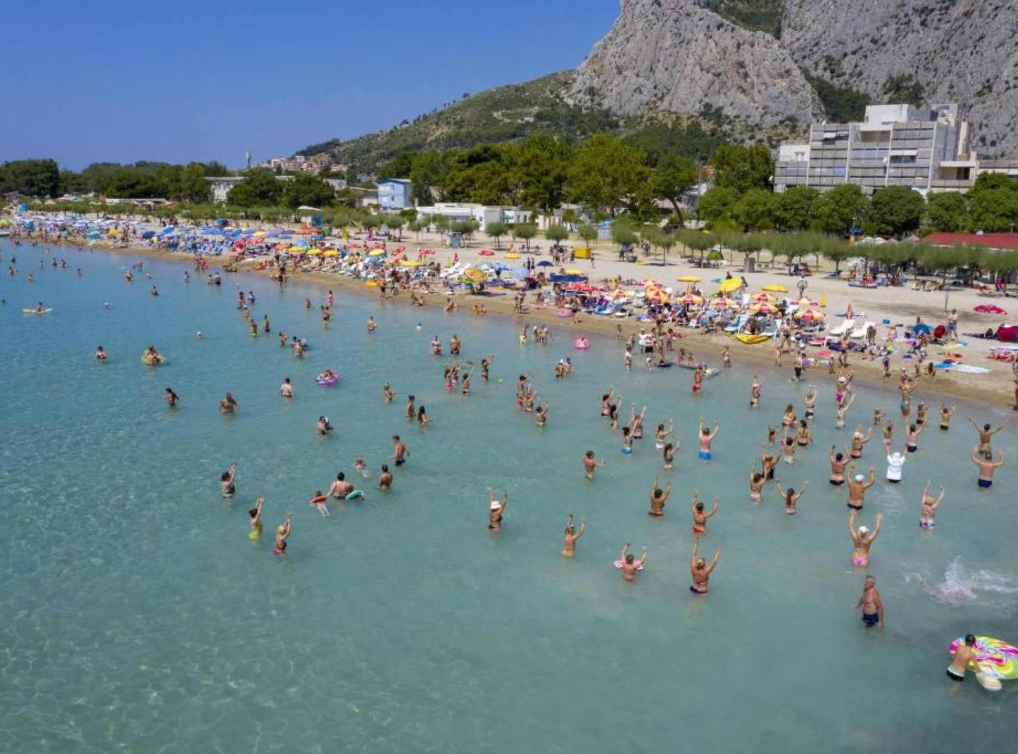 A crowded beach with clear water, umbrellas, and people enjoying the sun.