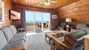 Ein Interieur einer Holzhütte mit Holzboden, Deckenventilator, Fernseher, Sofa, Tisch und Blick durch eine Glastür ins Freie.
