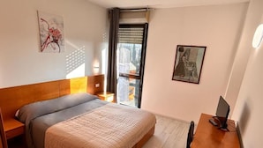 A bedroom with a bed, a desk, a chair, a TV, and a window with blinds.