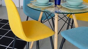 Uma sala de jantar com mesa redonda de vidro, cadeiras azuis e amarelas, e um revestimento de azulejos brancos ao fundo.
