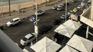 Uma estrada costeira com carros estacionados, palmeiras e praia ao fundo.