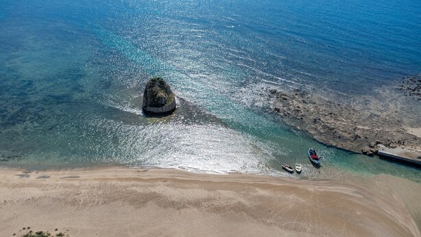 Vue aérienne d’une plage aux eaux turquoise et limpides, avec un promontoire rocheux et quelques bateaux près du rivage.