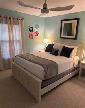 A bedroom with a bed, a ceiling fan, a nightstand, a lamp, and a window with curtains. - The Blue Retreat (Fort Myers Beach)