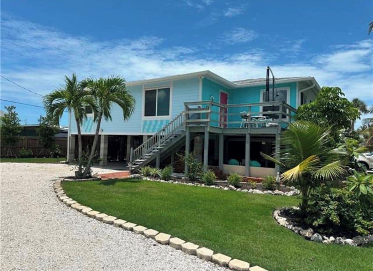 A two-story house with a balcony, a front porch, and a small garden with palm trees.