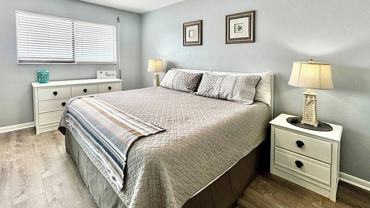 Une chambre à coucher avec un lit, des tables de chevet, une commode, une lampe et une fenêtre avec des stores.