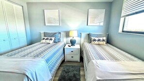 A bedroom with two beds, a nightstand with a lamp, and a window with blinds. - Beach Watch (Bonita Springs)