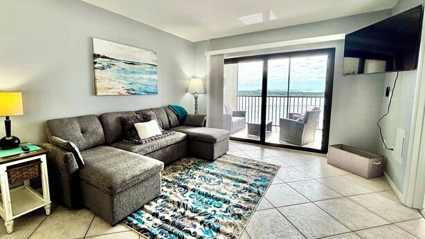 Een woonkamer met een grijze hoekbank, een kamerbreed tapijt, een schilderij van een strand en een balkon met uitzicht.