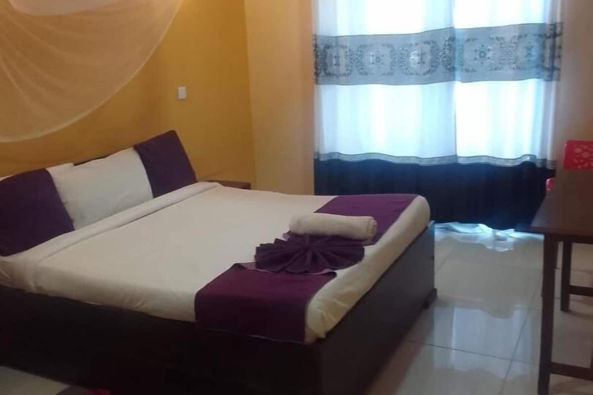 A neatly made bed with a white and purple bedspread, a dark wooden headboard, and a small table with a chair beside the bed.