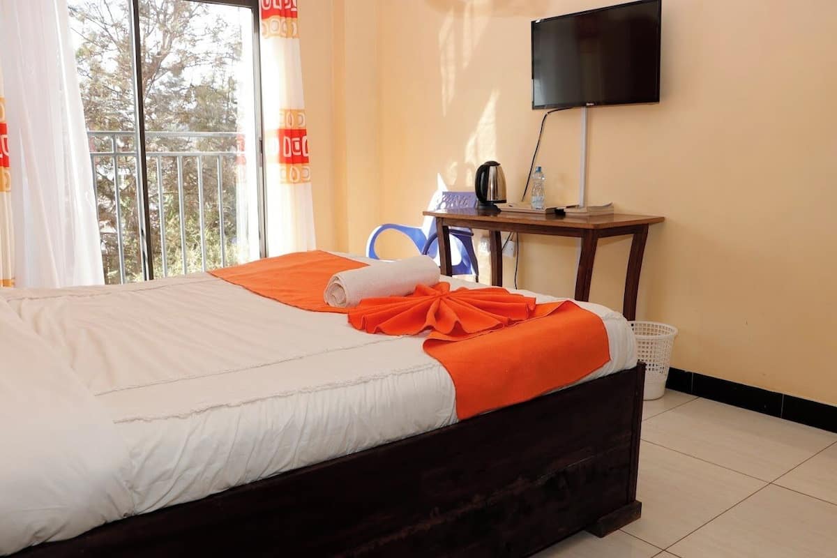 A hotel room with a bed, a television on a stand, a table with a kettle and a water bottle, and a window with curtains.