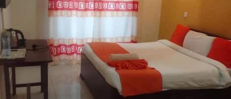 A hotel room with a bed, a desk, a chair, a table with a kettle, and a curtain.