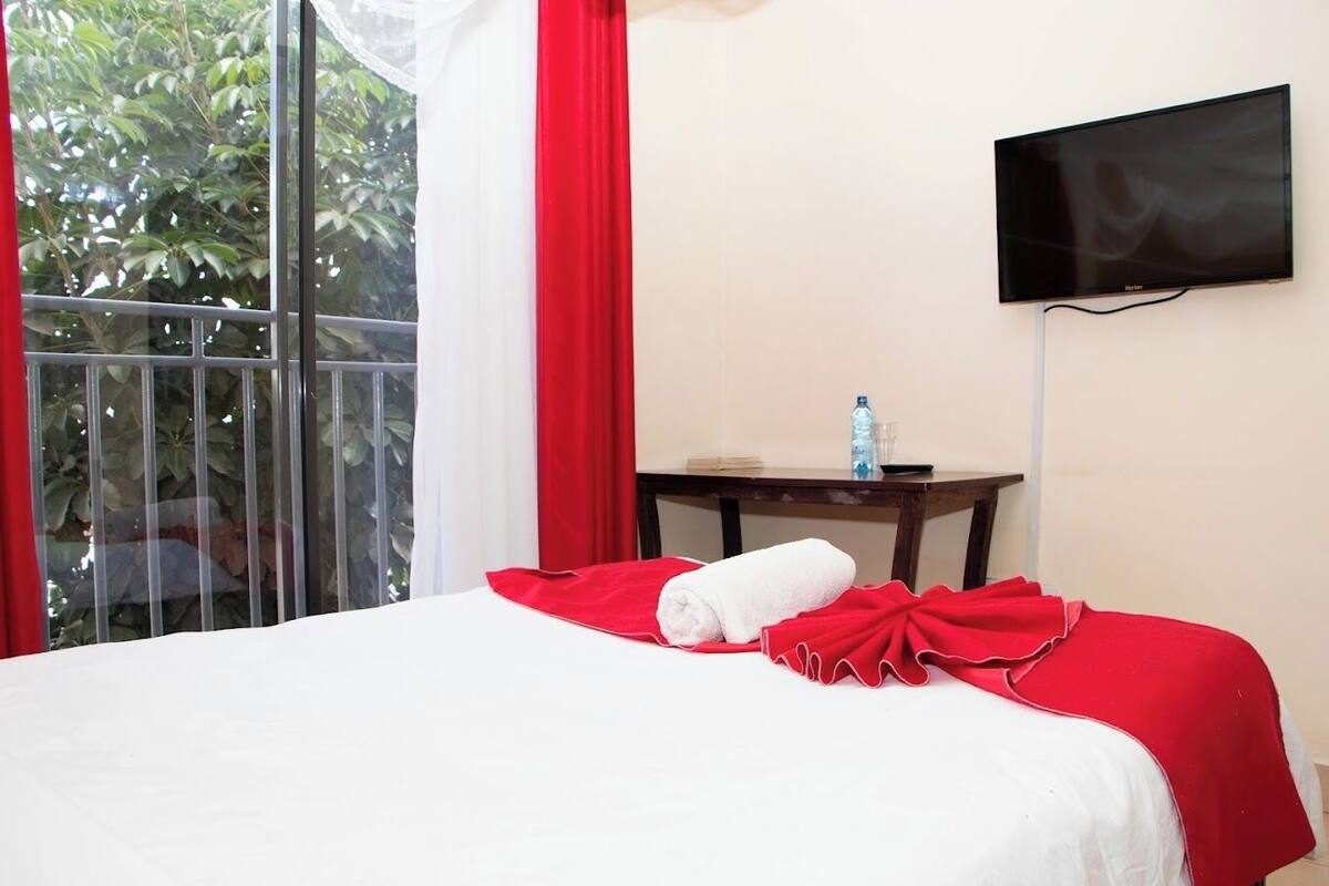 A hotel room with a bed, a TV mounted on the wall, a small table with a bottle and glass, and a view of greenery through a window.