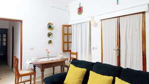 Un salon avec un canapé noir, des coussins jaunes, une table à manger en bois et un mur blanc orné d’une œuvre d’art colorée.