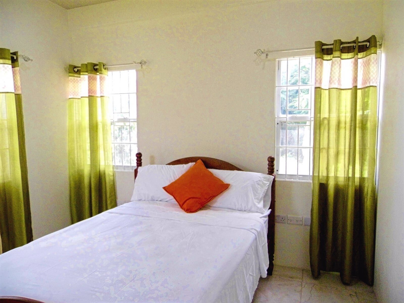 Un lit simple avec une literie blanche et un oreiller orange, encadré par deux fenêtres ornées de rideaux verts.