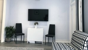Ein Wohnzimmer mit einem flachbildfernseher an der Wand, zwei schwarzen Sesseln, einem weißen Beistelltisch und einem gemusterten Sofa.