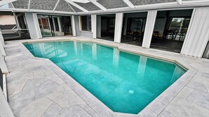 Ein rechteckiger Swimmingpool mit einer gefliesten Liegefläche, umgeben von einer überdachten Terrasse mit Glastüren zu einem Innenbereich.
