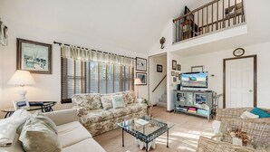 Un salon spacieux avec un canapé, une table basse en verre et un fauteuil en osier. Un escalier mène à un niveau supérieur, et il y a un téléviseur posé sur un meuble.