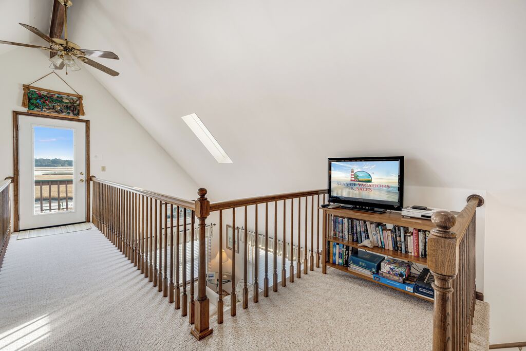 A spacious room with a sloped ceiling, a wooden staircase with a railing, a television on a stand, and a bookshelf filled with books.