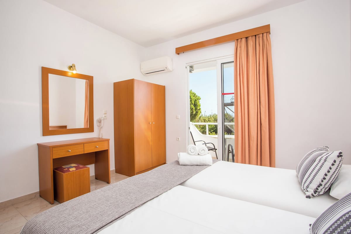 A hotel room with a bed, a wooden desk with a mirror, a wardrobe, and a sliding glass door to a balcony.