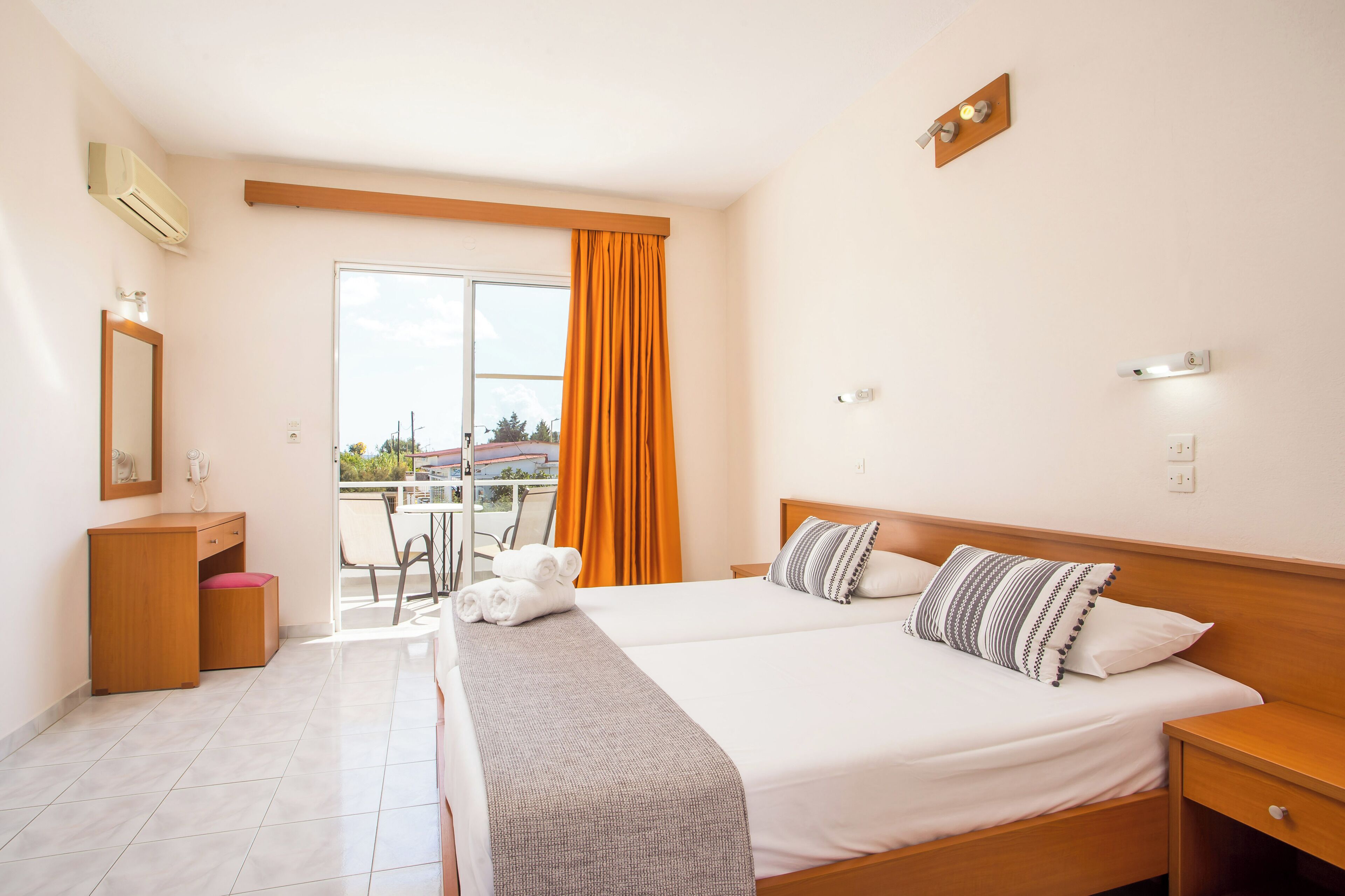 Ein Hotelzimmer mit zwei Betten, einem Schreibtisch, einem Stuhl und einem Balkon mit orangefarbenen Vorhängen.