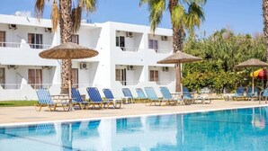 Zona de piscina amb gandules i ombrel·les, envoltada de palmeres i un edifici blanc amb balcons.