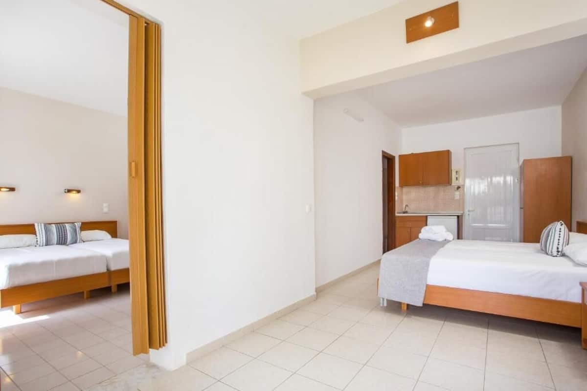 A hotel room with two beds, a wooden wardrobe, a small kitchenette, and a bathroom visible through an open door.