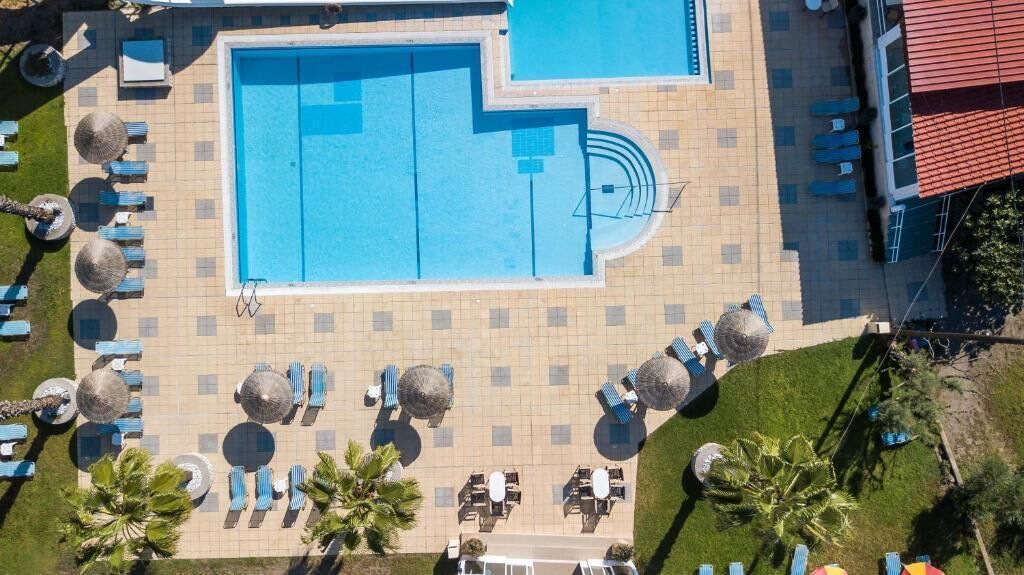 An aerial view of a swimming pool area with multiple lounge chairs and umbrellas, surrounded by a tiled deck and greenery.