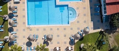 An aerial view of a swimming pool area with multiple lounge chairs and umbrellas, surrounded by a tiled deck and greenery.