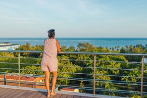 A person standing on a balcony overlooking a coastal view with trees and buildings in the background.