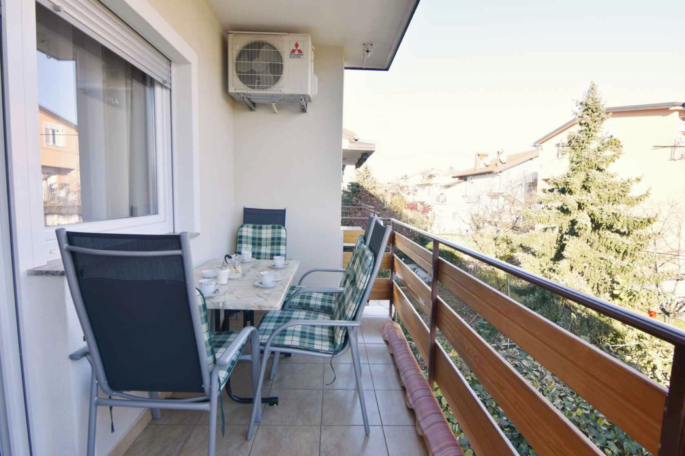 A balcony with a table and chairs, an air conditioning unit, and a view of the surrounding buildings and trees.
