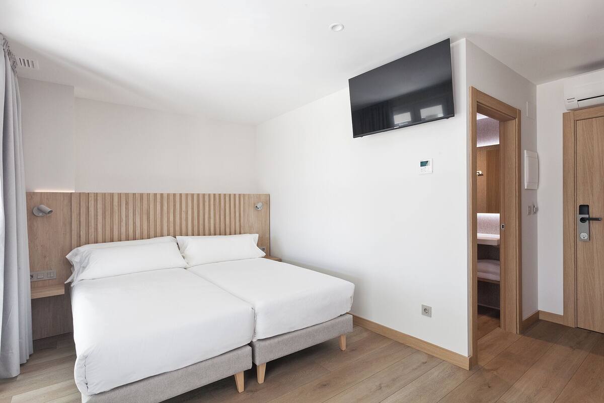 Habitación de hotel con una cama, un televisor montado en la pared, un cabecero de madera y una puerta que da a un baño.