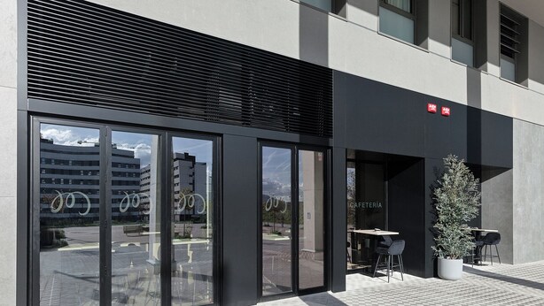A building entrance with large glass doors, a small outdoor seating area with tables and chairs, and a potted plant. - Hotel Iriguibel Arrosadía (Pamplona)
