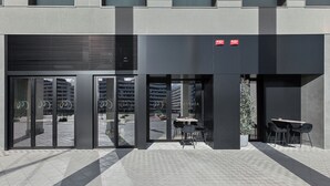 A modern building entrance with a glass door, a seating area with tables and chairs, and a potted plant. - Hotel Iriguibel Arrosadía (Pamplona)