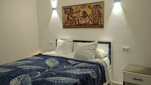 A bedroom with a bed, white pillows, a dark blue bedspread with leaf patterns, a wall-mounted painting, and two wall-mounted lights. - NOOR SEEN HOUSE  (Luxor)