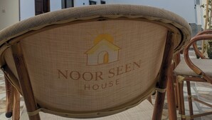 A chair with a woven backrest and a logo, positioned outdoors with a white building in the background. - NOOR SEEN HOUSE  (Luxor)