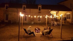 Ein Außenbereich mit Sitzgelegenheiten, Stühlen und einer Feuerstelle, umgeben von Lichterketten und einem Gebäude im Hintergrund.