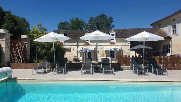 Ein Poolbereich mit Liegestühlen und Sonnenschirmen, im Hintergrund ein Steinhaus und ein klares, blaues Schwimmbecken.