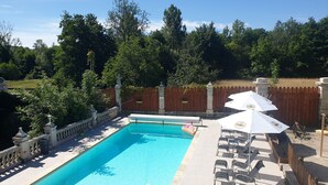 Ein rechteckiger Swimmingpool, umgeben von einem Holzzaun, mit Liegebereich, Stühlen und Sonnenschirmen sowie einer Steinbalustrade.