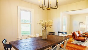 A dining area with a wooden table, white chairs, a vase with dried plants, and a leather sofa in the background.
