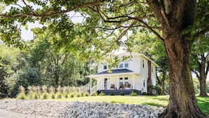 A white house with a porch, surrounded by a stone wall and trees.