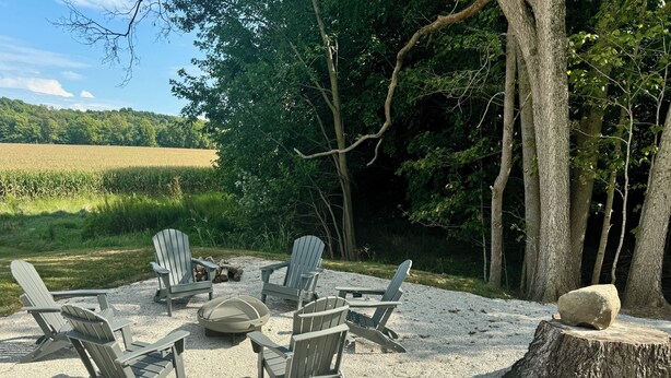 A gravel patio with Adirondack chairs and a fire pit, surrounded by trees and a field.
