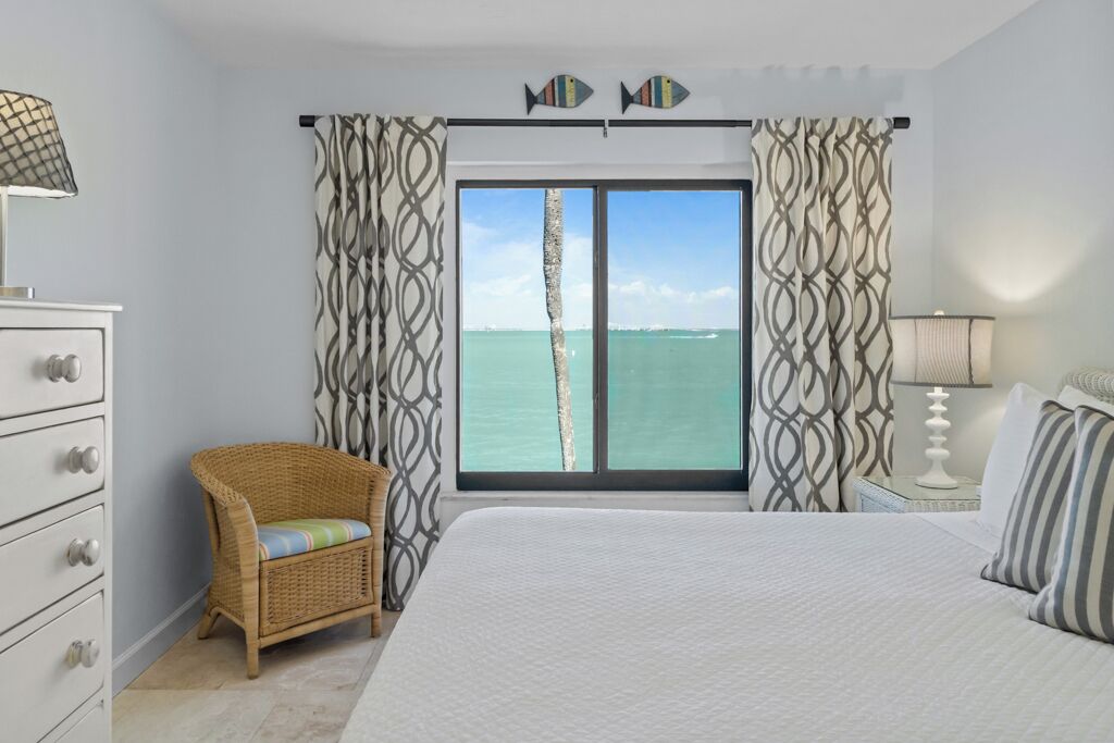 Quarto com uma janela grande, uma cama, uma cadeira de vime e um aparador.
