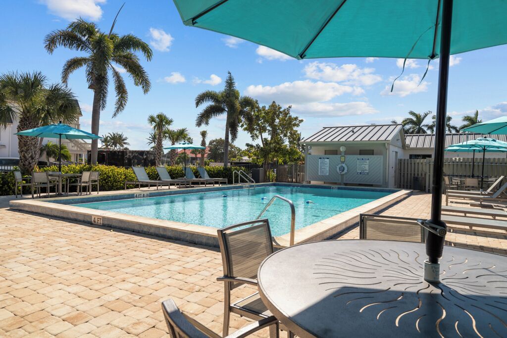 A pool area with lounge chairs and umbrellas, palm trees, and a building with a sign.