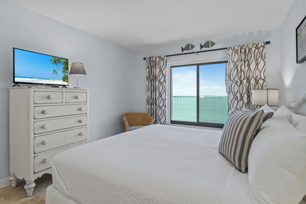 Une chambre avec un grand lit, un téléviseur posé sur une commode, un fauteuil et une vue sur la mer par la fenêtre.