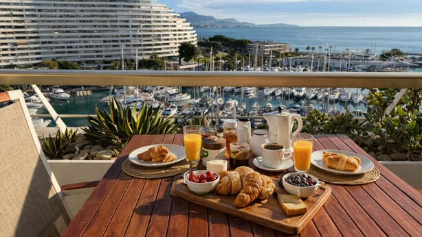 Ein Frühstückstisch mit Croissants, Beeren, Saft und Kaffee auf einem Balkon mit Blick auf einen Yachthafen und ein Kreuzfahrtschiff.